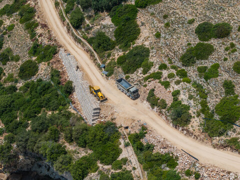 Aerial view of construction work on a dirt road featuring a yellow bulldozer and a dump truck alongside gabion retaining walls in the rocky landscape of Qeparo Fushe, Vlore County, Albania.