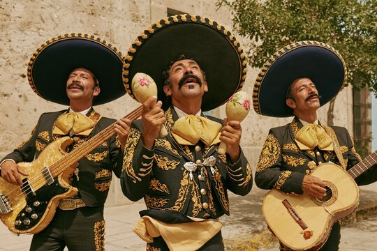 Three mariachi musicians in traditional charcoal embroidered suits perform on a city street, displaying weathered instruments and cultural pride for Mexican tourism and music content