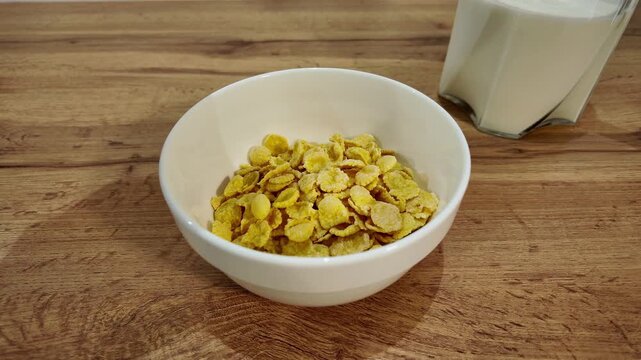 Yellow corn flakes falling into a white bowl on a wooden table. Healthy and nutritious meal preparation with a glass of milk nearby