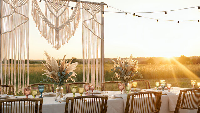 A long table is set up outdoors with colorful glasses and flower arrangements. String lights hang above as the sun sets behind distant hills. The scene captures a gathering atmosphere.