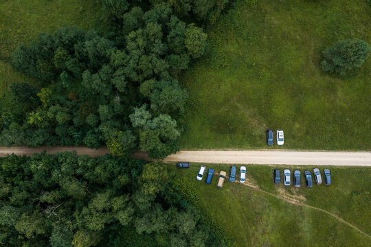 Aerial view of cars parked along a dirt road bordered by dense green forest and open grassy fields in Anyksciai, Utena County, Lithuania.