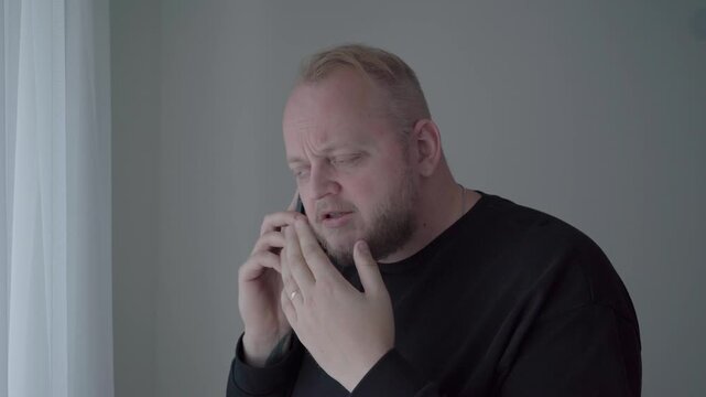Adult man by a bright window presses his ear and covers the other, clearly struggling to hear a muffled call through a poor connection.