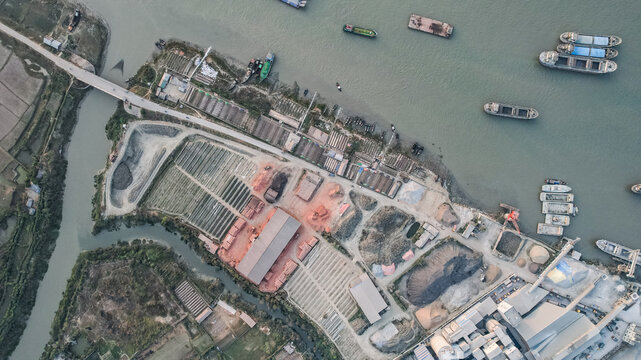 Aerial view of a brick kiln facility with rows of drying bricks, industrial structures, and cargo ships docked in the river in Chittagong, Chattogram, Bangladesh.