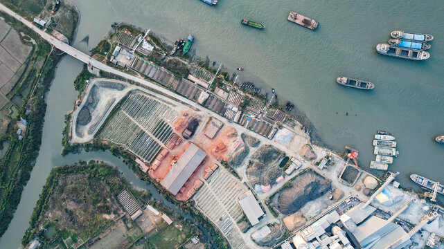 Aerial view of a brick kiln facility situated along the riverbank with cargo ships moored nearby, featuring rows of drying bricks and industrial structures in Chittagong, Chattogram, Bangladesh.