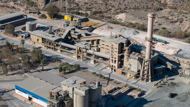 Aerial view of the Loma Negra cement plant with its large industrial kiln, tall smokestack, storage silos, and surrounding arid landscape in Rivadavia, San Juan Province, Argentina.