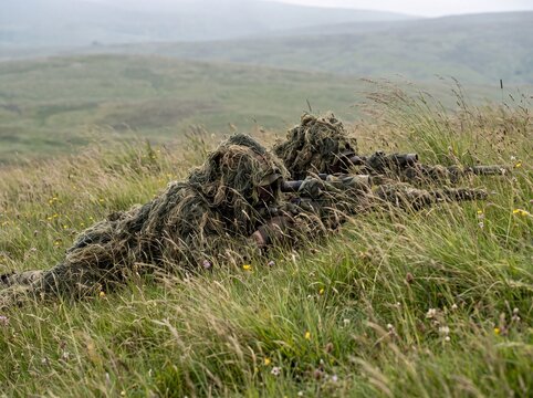 Camouflaged snipers in ghillie suits lying prone in tall grass on a misty hillside, observing their surroundings.