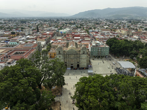 Aerial view of the Oaxaca Cathedral surrounded by historic colonial buildings, lush green trees in the zocalo, and the sprawling urban landscape of Oaxaca de Juarez, Oaxaca, Mexico.
