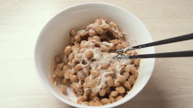 Chopsticks stirring fermented natto beans until sticky and stringy in white bowl, static shot