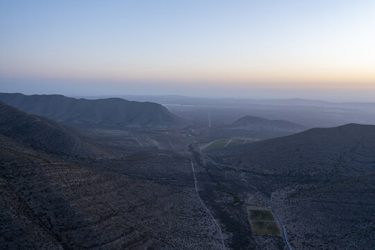 Aerial view of a valley featuring rows of vineyards nestled between rugged mountains under a soft twilight sky in Parras de la Fuente, Coahuila de Zaragoza, Mexico.
