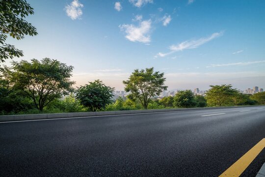 Empty asphalt highway with white lane markings stretches toward urban skyline. Modern city buildings, skyscrapers appear under blue sky with clouds. Green trees line roadside near empty street in