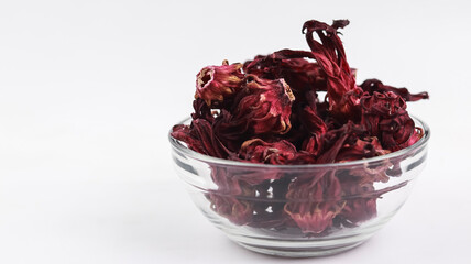 A close-up, side-angle view of a glass bowl filled with dried dark red roselle hibiscus flowers, positioned against a clean white background with copy space on the left. © VH_World