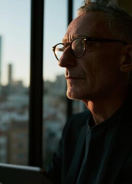 Portrait of a middle-aged man with short grey hair and glasses. he is looking out of a window with a view of a city skyline in the background.