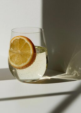 Glass of white wine with a slice of orange on the rim. the glass is placed on a white surface with a shadow cast on the right side of the image. the background is a plain white wall.