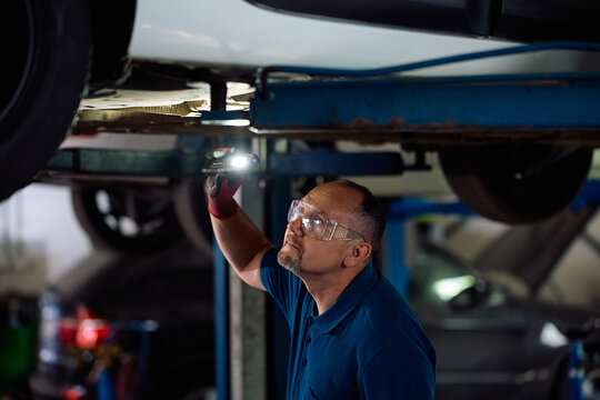 Car mechanic using flashlight while checking undercarriage in repair shop.
