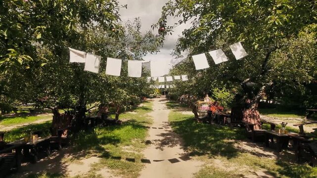 Outdoor cafe patio with bistro tables under apple trees and white fabric garland against cloudy sky. Rustic garden terrace with wooden bar, folding chairs and stone brick patio in summer orchard