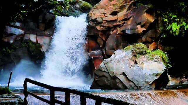 A view of a natural waterfall cascading between sturdy rock cliffs with reddish and brownish hues.