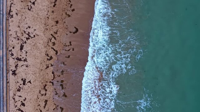 Drone flies directly above Longchamp Beach, Saint-Lunaire, Ille-et-Vilaine, Brittany, France, revealing breaking waves, sandy shoreline, seaweed deposits and the English Channel in a top-down forward