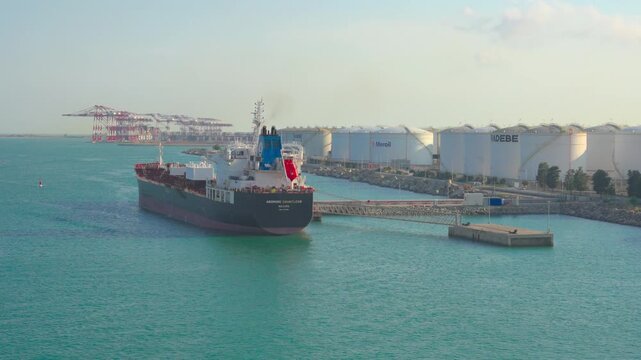 Large freight ship slowly turning in the calm waters of an industrial harbor, with oil storage tanks and gantry cranes visible on the shoreline under a clear sky during daytime