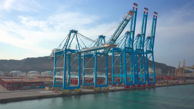Large blue gantry cranes operating at a container terminal, moving along the quay for loading and unloading cargo ships, with shipping containers stacked in the background logistics area