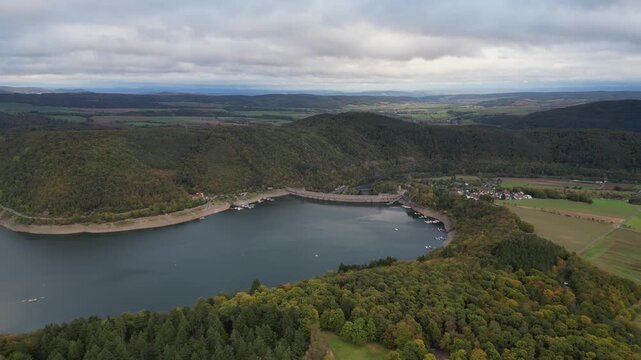 Drone view over the expansive Edersee reservoir and its impressive dam, nestled in the lush, hilly landscape of Hesse, Germany. A tranquil setting with a small village and boats on the water.