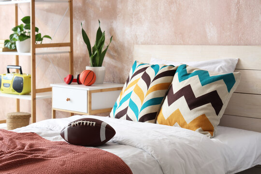 American football on a bed with patterned pillows and a nightstand in a modern bedroom interior, an american football rests on a bed with a white duvet and a rust-colored knitted blanket