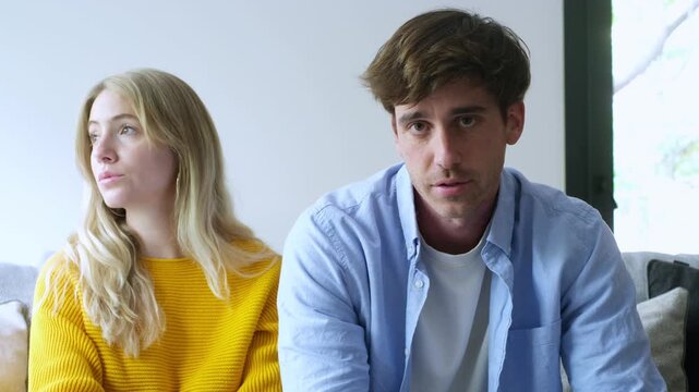 A young couple sits on a couch, looking at the camera with serious expressions. Their faces convey deep thought, concern, or perhaps a moment of shared reflection in their modern living room.