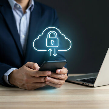 Businessman holds smartphone displaying cloud security pad lock icon near laptop on office desk.
