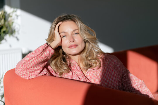 Mature woman rests her head in one hand on an orange sofa in sunlight, physical fatigue, low energy, sick day, home recovery.