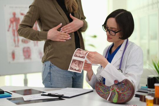 Woman doctor shows abdominal ultrasound pictures to standing female patient in clinic. Focused specialist points at scan explaining liver pain concern