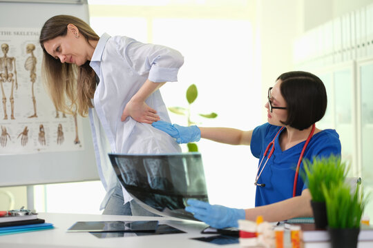 Woman doctor checks female lower back pain during orthopedic clinic visit. Focused medic points to sore spine reviewing X-ray scan with patient