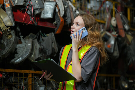 Warehouse Manager: Determined young woman, wearing safety vest and holding a clipboard, speaks on her phone amidst shelves filled with automotive parts. Scrap Yard recycle auto engine workplace