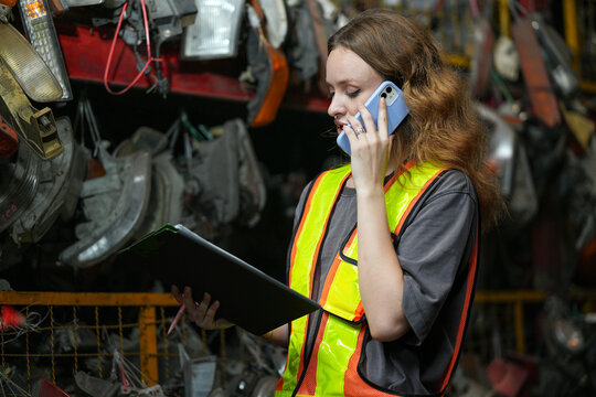 Warehouse Manager: Determined young woman, wearing safety vest and holding a clipboard, speaks on her phone amidst shelves filled with automotive parts. Scrap Yard recycle auto engine workplace