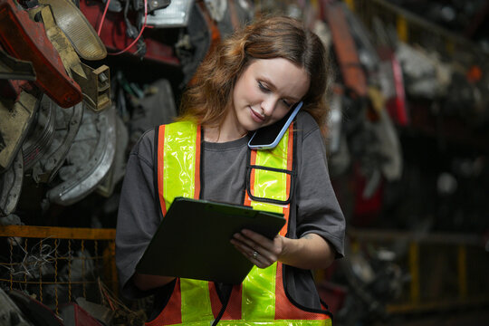Warehouse Manager: Determined young woman, wearing safety vest and holding a clipboard, speaks on her phone amidst shelves filled with automotive parts. Scrap Yard recycle auto engine workplace