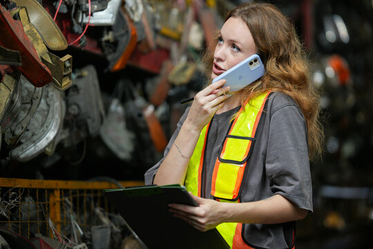 Warehouse Manager: Determined young woman, wearing safety vest and holding a clipboard, speaks on her phone amidst shelves filled with automotive parts. Scrap Yard recycle auto engine workplace