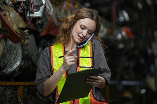 Warehouse Manager: Determined young woman, wearing safety vest and holding a clipboard, speaks on her phone amidst shelves filled with automotive parts. Scrap Yard recycle auto engine workplace
