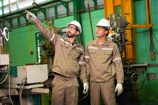 Engineer factory worker helmet machine safety industrial workshop pointing team discuss production task near heavy equipment, protective uniform, glove, glasses, green wall, indoor plant setting