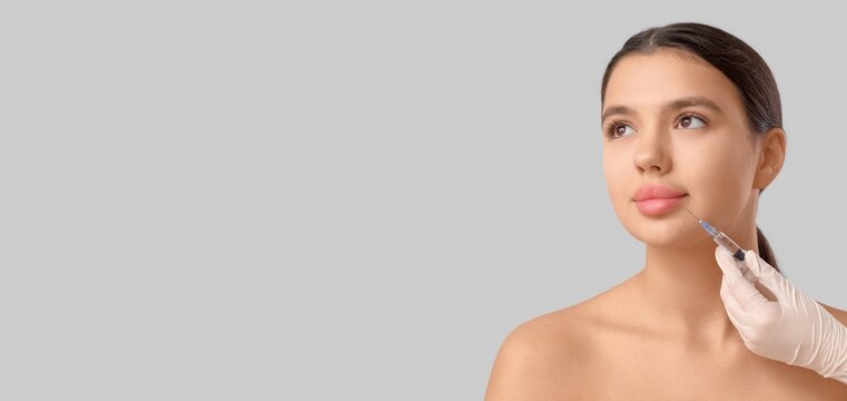 Young woman receiving lip filler injection, cosmetic procedure for beauty and anti-aging, isolated on a gray background