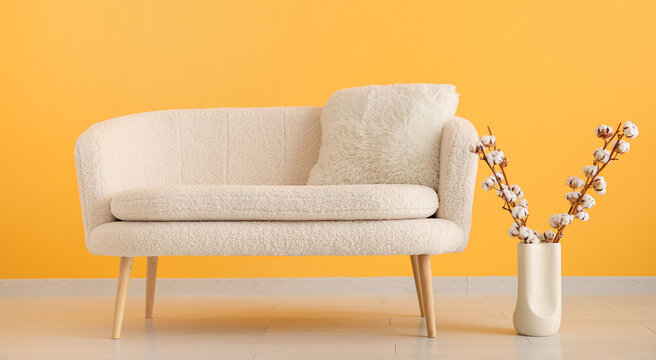 Cozy cream boucle sofa with a fluffy pillow and cotton branches in a vase against a bright yellow wall, modern home interior design