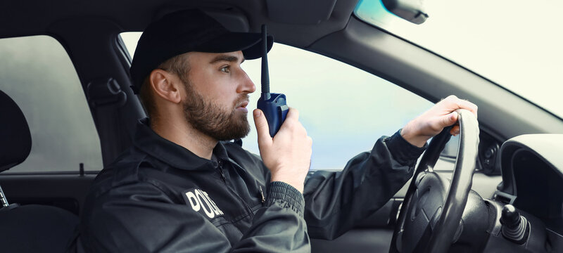 Male police officer in uniform driving a patrol car and communicating with a walkie-talkie, He is holding a walkie-talkie to his mouth