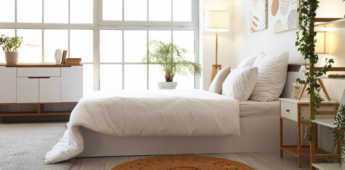 Fototapeta premium Bright and airy bedroom interior with white bedding, large window, green plants, and modern furniture for home decor
