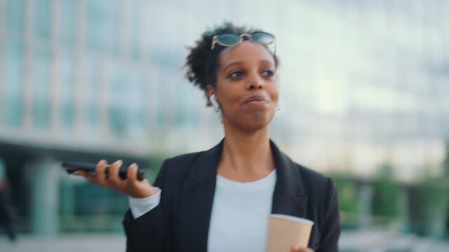 Portrait of contemporary business woman walking in city street, talk by mobile. Joyful black woman holding coffee cup in hand, listening to sounds in wireless earphone, mobile call to colleague