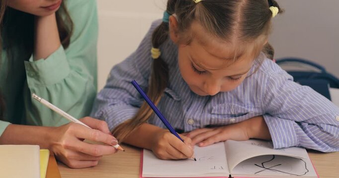 A young mother helping her little girl write and draw in a notebook with pencils at home.
