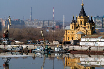 Obraz premium A yellow Orthodox cathedral on an industrial embankment with cranes, boats and a reflection of the cityscape in Nizhny Novgorod, Russia.