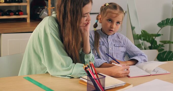 Mother guiding and helping young daughter writing homework in notebook at study desk.