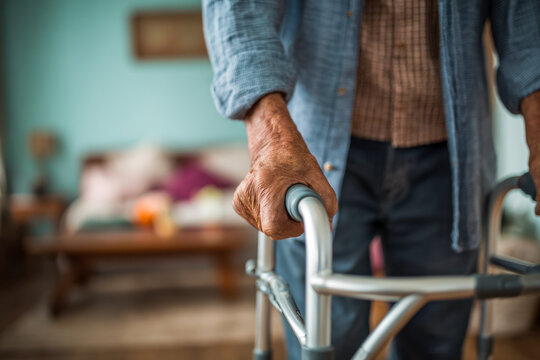 Elderly person using a walker for support while standing indoors with blurred home surroundings in the background showing signs of assisted mobility and care