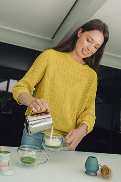 Smiling woman pours milk into a glass of matcha tea in the kitchen