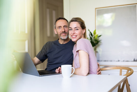 Mature couple embraces while sitting at a table with a laptop