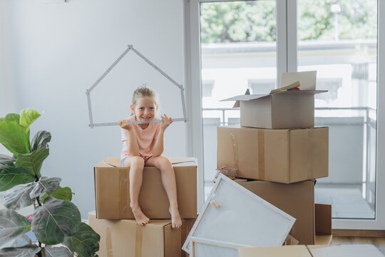 Child sitting on moving boxes holding house shape indoors on moving day
