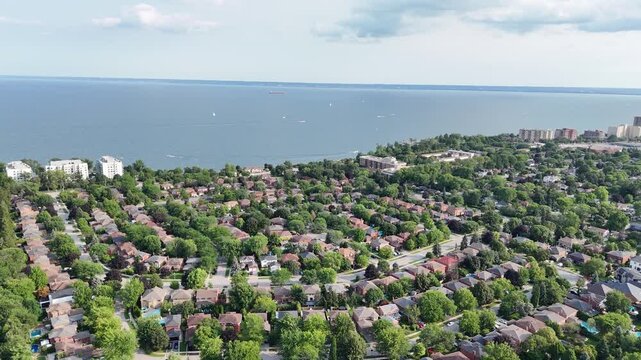 Wide aerial tracking shot flying forward over a dense suburban neighborhood of detached houses and lush green tree canopies toward the open blue waters of Lake Ontario in Oakville, Canada