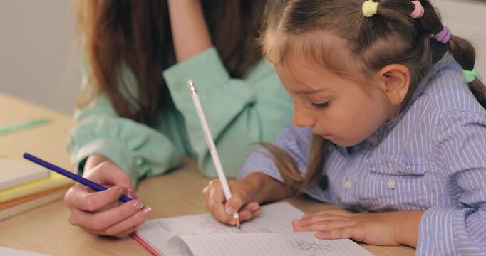A young mother helping her little girl write and draw in a notebook with pencils at home.